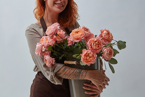 Smiling girl in a brown apron with a tattoo holds a beautiful bouquet of pink roses in a vase on a gray background with space for text. The concept of Valentine's Day.