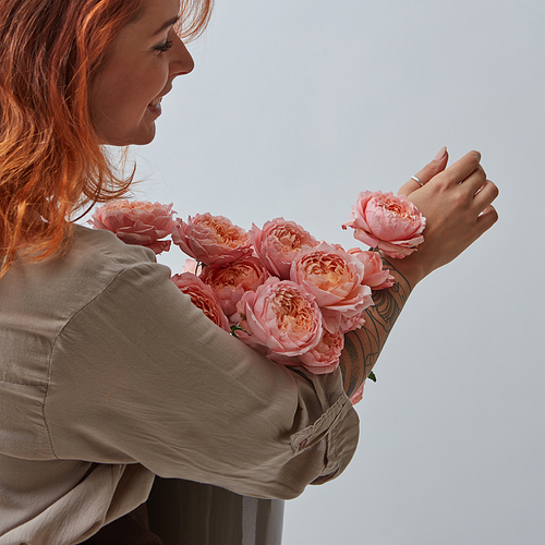 Delicate pink bouquet of roses in a vase holds in hands a pretty red-haired woman with a tattoo around a gray background with space for text. Mothers Day