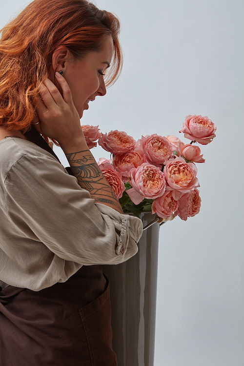 Girl florist with a tattoo in a brown apron holding a bouquet of pink roses in a vase around a gray background with space for text. The concept of a flower shop