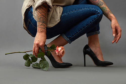 Pink flower in a girl's hand with a tattoo squatting. Young stylish girl in high-heeled shoes, and jeans on a dark background with copy space.