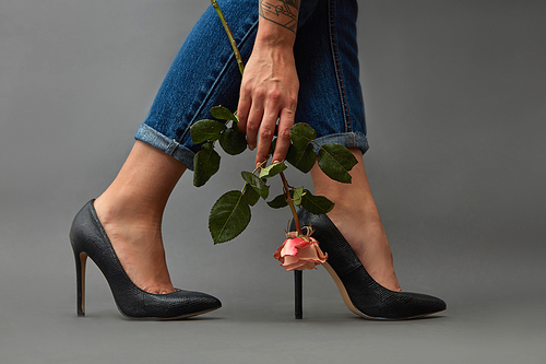 Girl with a tattoo in jeans, and black heeled shoes, holding a fresh pink flower around a dark background with space for text. Card