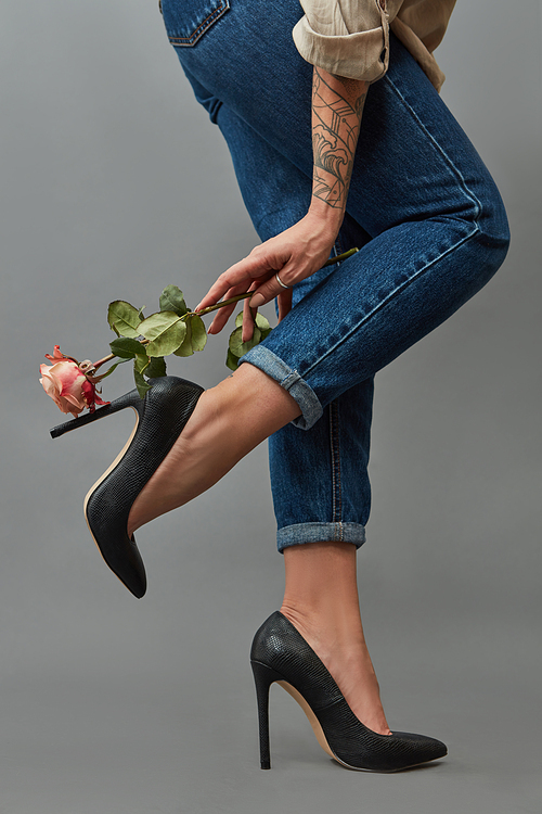 A woman's hand with a tattoo is holding a pink rose near her legs in jeans and stylish high-heeled shoes around a dark background with space for text. Photo as layout for a postcard