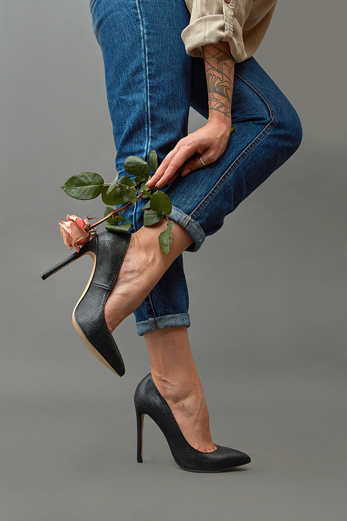Legs of a girl in classic black high-heeled shoes and jeans near a female hand holding an elegant pink flower around a dark background with copy space. Stylish composition