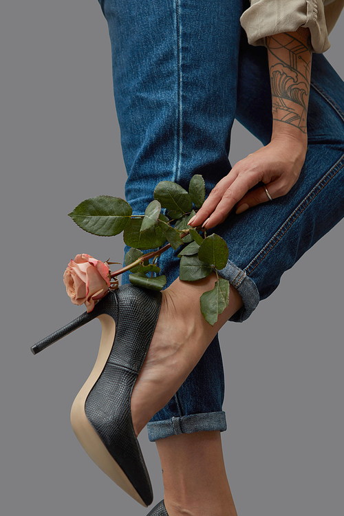 Hand of a girl with a pink flower beautifies a leg in jeans and stylish black high-heeled shoes around a dark background with copy space.
