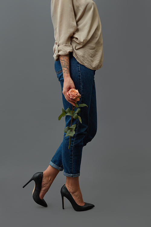 Elegant fashionable girl in jeans, black high-heeled shoes and with a tattoo is holding a fresh rose flower around a dark background with copy space for text.