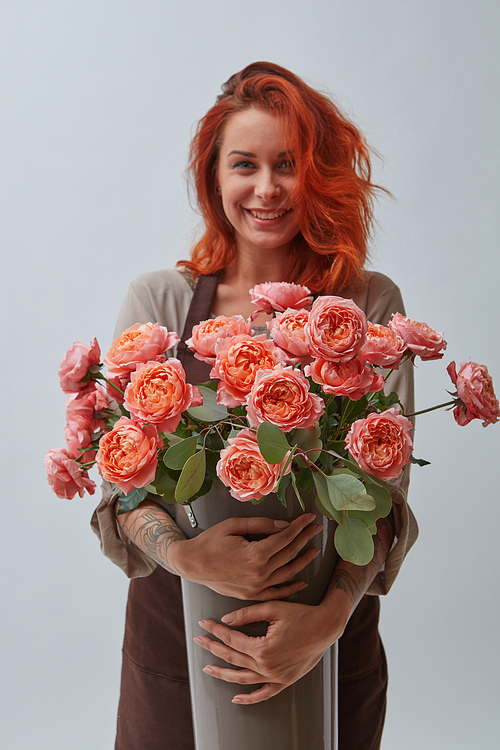 A smiling red-haired woman in a brown apron holds a vase with roses in a color of the year 2019 Living Coral Pantone on a gray background with space for text. The concept of a flower shop