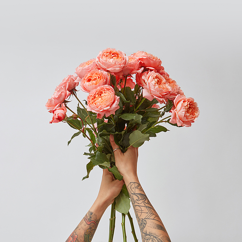 Beautiful bouquet of fresh roses in woman's hands with tatoo in a color of the year 2019 Living Coral Pantone on a gray background with copy space. Top view.