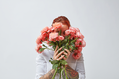 A happy woman with a tattoo on her hands hides her face behind a bouquet of fresh roses in a color of the year 2019 Living Coral Pantone. Postcards for Mother's Day.