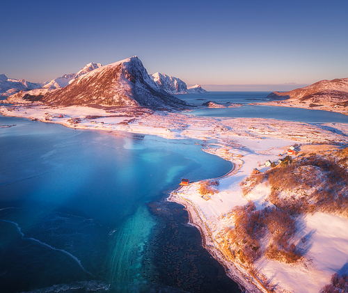 Aerial view of snow covered mountains, houses, clear water and blue sky at sunset. Winter landscape with sea coast, snowy rocks, rorbu, road. Top view of small village in Lofoten islands, Norway
