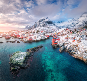 Aerial view of small village at sunset in winter. Travel in Lofoten islands, Norway. Landscape with blue sea, snowy mountains, high rocks, buildings, traditional rorbu, cloudy sky. View from above