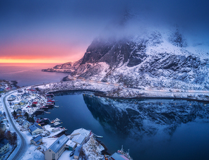 Aerial view of snowy mountain, village on sea coast, colorful sky at sunset in winter. Top view of Reine, Lofoten islands, Norway. Moody landscape with high rocks, houses, rorbu, reflection in water