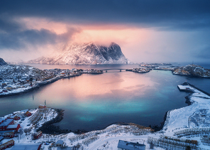 Aerial view of snowy mountain, village on sea coast, orange sky at sunset in winter. Top view of Reine, Lofoten islands, Norway. Moody landscape with high rocks, houses, rorbu, reflection in water
