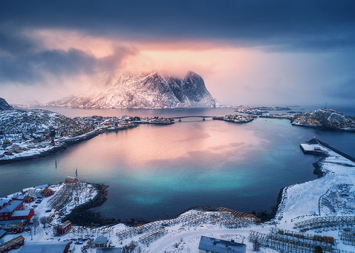 Aerial view of snowy mountain, village on sea coast, orange sky at sunset in winter. Top view of Reine, Lofoten islands, Norway. Moody landscape with high rocks, houses, rorbu, reflection in water