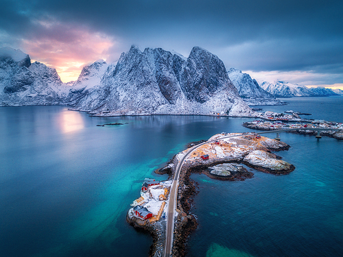 Aerial view of Hamnoy at dramatic sunset in winter in Lofoten islands, Norway. Moody landscape with blue sea, snowy mountains, rocks, village, buildings, rorbu, road, bridge, cloudy sky. Top view