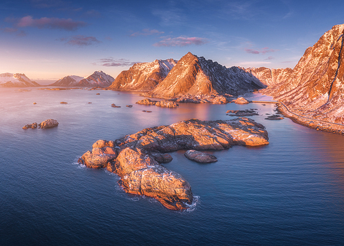 Aerial view of rocks in the sea, snowy mountains, blue sky with clouds at sunset. Travel in Lofoten Islands, Norway. Winter landscape with small islands in water, cliffs and waves. View from above
