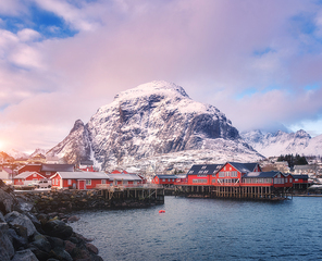 Red traditional rorbuer in small village at sunset in winter in Lofoten islands, Norway. Landscape with sea, snowy mountains, houses, blue sky with purple clouds. Beautiful rorbu on sea coast. Travel