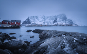 Beach with stones in blurred water, red rorbu and snowy mountains in fog at dusk in winter. Sea coast in Lofoten islands, Norway. Dramatic landscape with houses, sea. Moody scenery. Norwegian rorbuer
