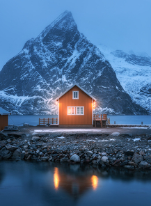 Red rorbu on sea coast and snow covered mountain at night. Lofoten islands, Norway. Moody winter landscape with traditional norwegian rorbuer, reflection in water, snowy rocks. Old fishermen house