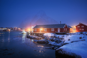 Beautiful norwegian Rorbu on sea cost and snowy mountain at night in winter. Lofoten islands, Norway. Landscape with red houses, fishing boats, snow covered rocks  in fog, reflection in water at dusk