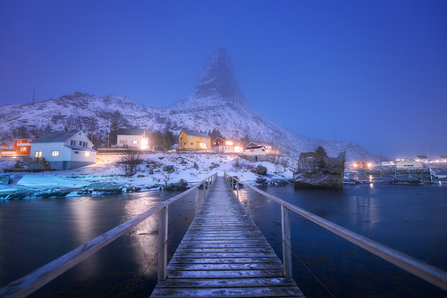 Wooden bridge, houses and rorbu on sea coast and snowy mountain in fog at night in winter in Reine village, Lofoten islands, Norway. Beautiful landscape with buildings, city illumination, high rocks