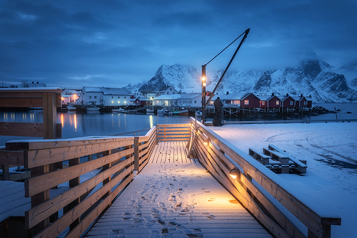 Snowy wooden pier on the sea coast with lights, rorbu and houses, boats and snow covered mountains in clouds at night. Landscape with jetty, buildings, rock in fishing village. Lofoten islands, Norway
