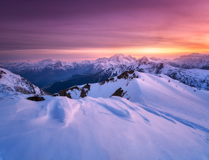Colorful red sky with clouds and bright sunlight over the snow covered mountains at sunset in winter. Beautiful wintry landscape with snowy rocks and hills at dusk. Scenery with alps at frosty evening