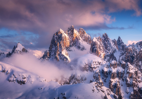 Majestic snowy mountain peaks in fog  and blue sky with purple clouds at sunset. Winter landscape with beautiful snow covered rocks in low clouds in frosty evening.  Nature in Dolomites, Italy. Alps