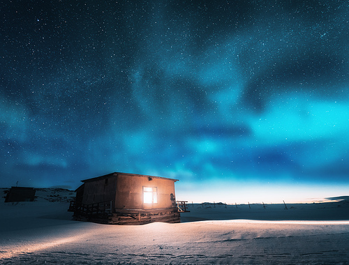 Aurora borealis over old small house with light from the window in winter. Northern lights in Teriberka, Russia.  Blue starry sky and green polar lights. Night landscape with aurora and building