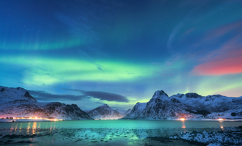 Aurora borealis over the sea coast, snowy mountains and city lights at night. Northern lights in Lofoten islands, Norway. Starry sky with polar lights. Winter landscape with aurora reflected in water