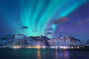 Aurora borealis over the sea, snowy mountains and city lights at night. Northern lights in Lofoten islands, Norway. Purple starry sky with polar lights. Winter landscape with aurora reflected in water