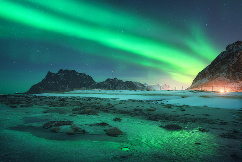 Aurora borealis above the snowy mountain and sandy beach in winter. Northern lights in Lofoten islands, Norway. Starry sky with polar lights. Night landscape with aurora, frozen sea coast, city lights