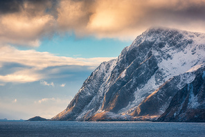 beautiful high snow covered mountains at  in winter. lofoten islands, norway. landscape with snowy rocks, sea, blue sky with orange clouds in the evening. travel and nature