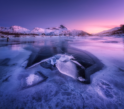 Snowy mountains, blue sea with frosty coast, reflection in water and purple sky at colorful sunset in Lofoten islands, Norway. Winter landscape with snow covered rocks, fjord with ice at night. Nature