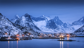 Houses in village, city lights, snowy mountains, sea, blue cloudy sky reflected in water at night in winter. Landscape with beautiful Reine at night in Lofoten islands, Norway. Norwegian rorbu at dusk