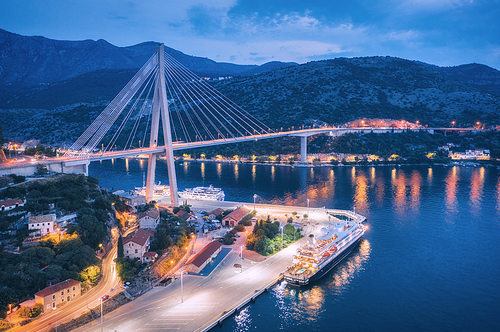 Aerial view of cruise ship in port and beautiful bridge at night. Landscape with ships and boats in harbour, city lights, road, mountains, blue sea at sunset. Top view. Floating liner at harbor