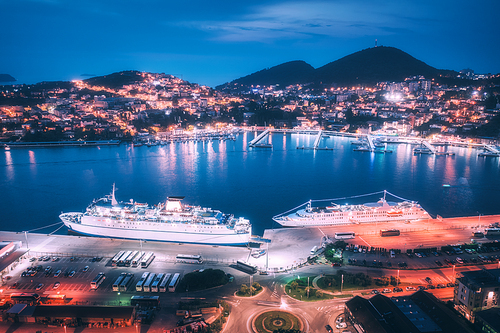 Aerial view of cruise ship in port at night. Landscape with ships and boats in harbour, city lights, buildings, mountains, blue sea at blue hour. Top view. Luxury cruise. Floating liner at harbor