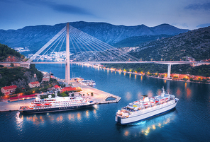 Aerial view of cruise ship at harbor and beautiful bridge at night. Landscape with ships and boats in harbour, city lights, road, mountain, blue sea at sunset. Top view. Floating liner in port at dusk