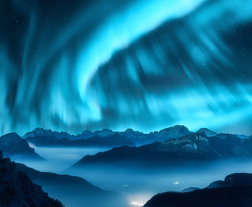 Aurora borealis above the mountains in fog at night. Northern lights. Sky with stars with polar lights and high rocks. Beautiful landscape with aurora, city lights in low clouds, mountain peaks. Space