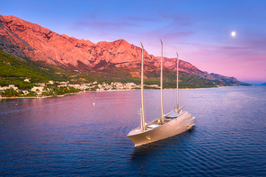 Luxury yacht and blue sea at sunset in summer. Aerial view of big modern sail boat. Top view of beautiful futuristic yacht, water, city, green trees, mountains, purple sky with moon. Adriatic sea