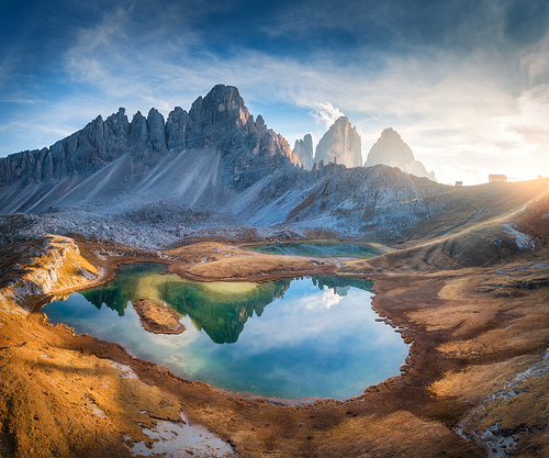 Aerial view of beautiful rocks, mountain lake, reflection in water and houses on the hill at sunset. Autumn landscape with mountains, blue sky and sunlight. Dolomites, Italy. Top view of Italian alps