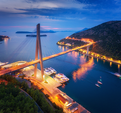Aerial view of beautiful modern bridge at night. Dubrovnik, Croatia. Top view of road, boats, yachts and city lights at sunset. Summer landscape with harbor, mountain, highway, blue sea and cloudy sky