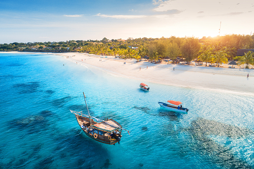 Aerial view of the fishing boats on tropical sea coast with sandy beach at sunset. Summer holiday in Zanzibar, Africa. Landscape with boat, yacht in transparent blue water, green palm trees. Top view