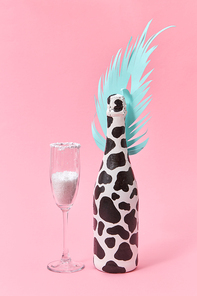 Holiday decorative composition of painted champagne bottle with black spots and paper cut tropical leaf with glass of white powder on a pastel millennial pink background, copy space. Holiday card.