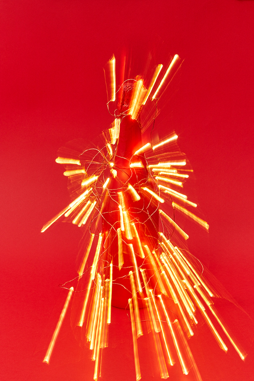 Firecracker from shined trails of Christmas lights on a painted wine bottle on a red background with copy space. New Year congratulation card.