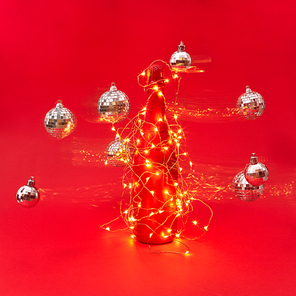 Creative composition from painted champagne bottle covered Christmas garland with shining lights and glitter balls floating around with trails on a red background, copy space. New Year greeting card.