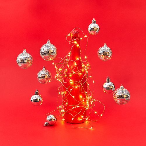 New Year shined lights string on a creative painted wine bottle and floating balls around on a red background with copy space. Christmas congratulation card.