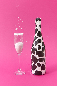 Decorative white painted wine bottle with black spots and glass with falling white powder on a hot pink background, place for text. Congratulation card.