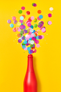 Holiday champagne red painted bottle with multicolored glitter as a bubbles foam on an yellow background, copy space. Flat lay.