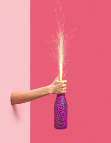 Woman's hand holds painted champagne bottle with holiday firecracker on a duotone pink background, copy spase. Christmas concept.