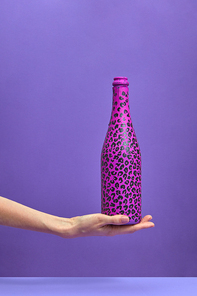 The champagne bottle painted hot pink and black spots on a woman's hand on a lilac background, copy space. Flat lay. Holiday concept.